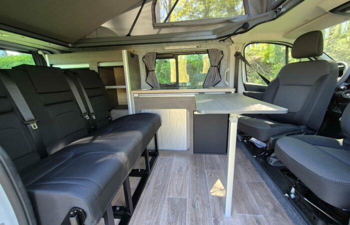 Intérieur d'un camping-car avec sièges noirs, table pliante, plancher imitation bois, kitchenette avec évier et grandes fenêtres avec rideaux. Le toit surélevé offre davantage d'espace pour la tête et la lumière naturelle.