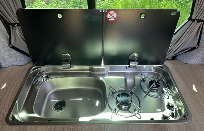 Une cuisine compacte en acier inoxydable pour camping-car, avec un évier à gauche et deux feux à gaz à droite, recouverte d'un couvercle noir à charnière orné d'un panneau « Interdiction de fumer ». La verdure est visible par la fenêtre derrière.