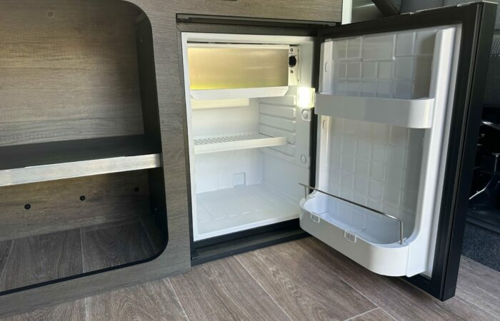 Un petit réfrigérateur vide, la porte ouverte, est encastré dans un meuble moderne lambrissé. Propre et éclairé, il laisse apparaître des étagères et des compartiments de rangement sur la porte. Le parquet marron est visible.