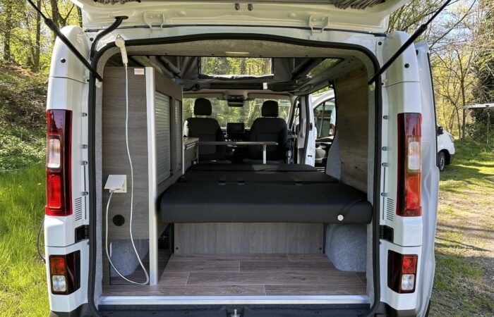 L'image montre l'intérieur d'un camping-car vu depuis les portes arrière ouvertes. Le camping-car est équipé d'un lit pliant, d'un plancher imitation bois, de sièges à l'avant et d'une lumière naturelle provenant des portes et fenêtres ouvertes.