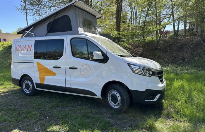 Un camping-car blanc à toit relevable est garé sur l'herbe, près des arbres. Il arbore des graphismes orange et le mot « OVAN » sur le flanc. La lumière du soleil filtre à travers les arbres, créant un paysage extérieur paisible.