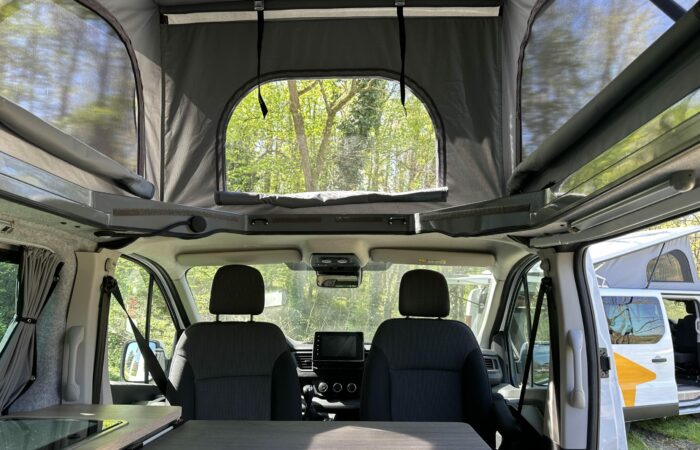 Vue intérieure d'un camping-car équipé de deux sièges avant, d'une table et d'un toit relevable surélevé avec fenêtres grillagées, laissant entrer la lumière naturelle et offrant une vue sur les arbres. Un autre camping-car est partiellement visible par la porte ouverte.