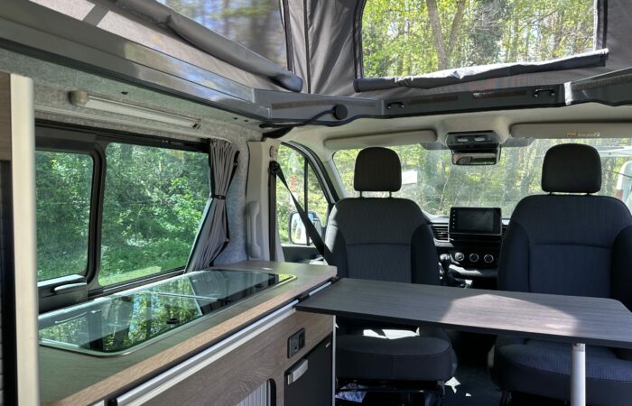Intérieur d'un camping-car avec deux sièges avant, un toit relevable, une table pliante et une kitchenette compacte près de la fenêtre. La lumière du soleil filtre à travers les arbres, créant une atmosphère lumineuse et chaleureuse.