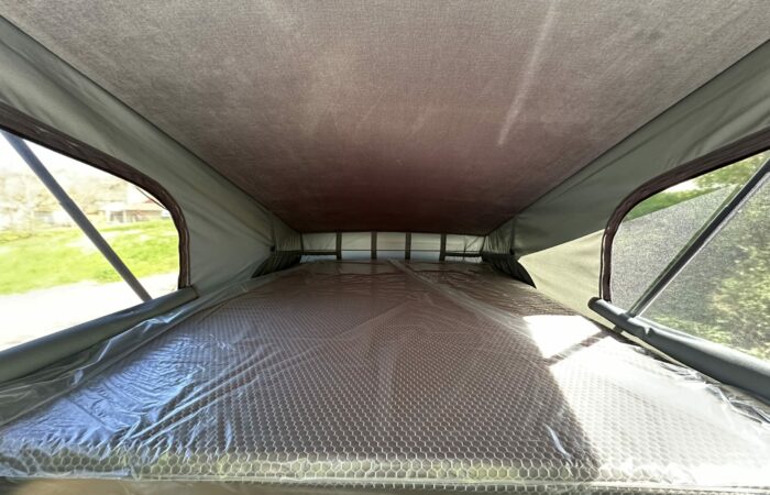 Vue intérieure d'une tente de toit avec un matelas recouvert de plastique, des fenêtres en filet des deux côtés et la lumière du soleil qui pénètre, créant un environnement lumineux et spacieux pour le camping.