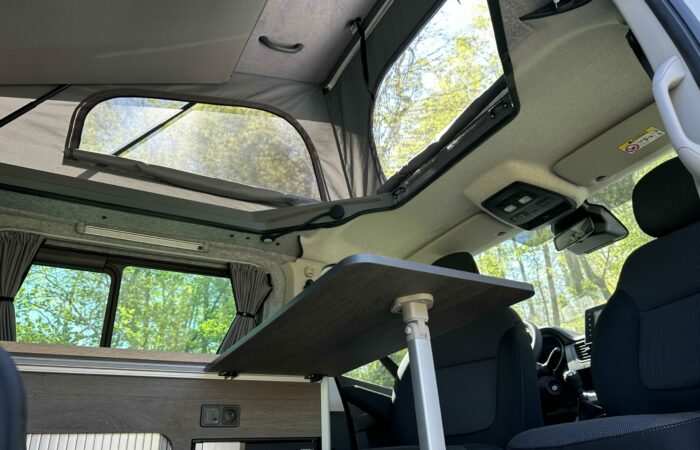 Intérieur d'un camping-car avec un toit relevable surélevé, une table en bois, des sièges noirs et la lumière du soleil entrant par les fenêtres ouvertes, révélant une vue sur les arbres à l'extérieur.